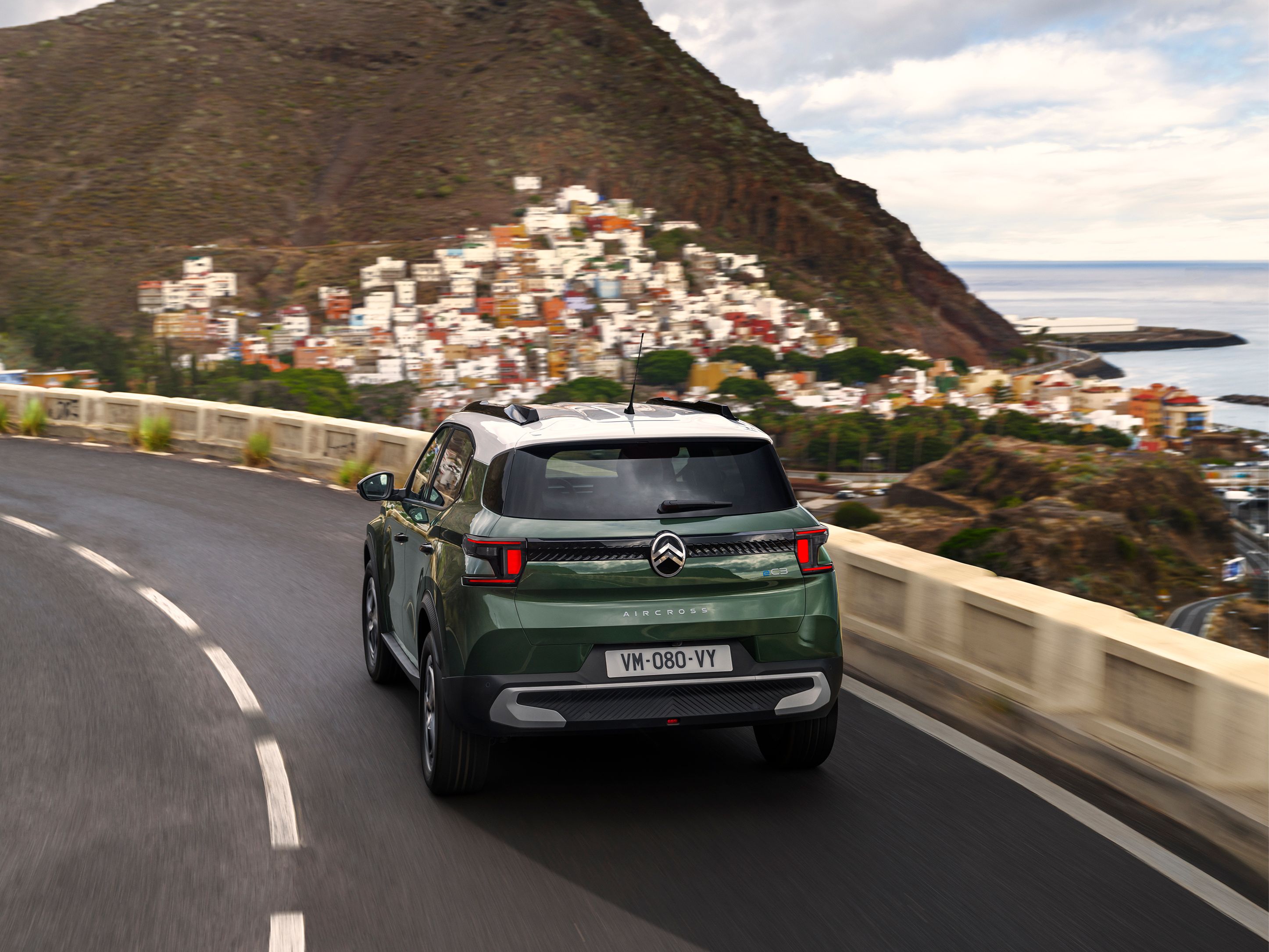 Vue arrière trois quarts du SUV Citroën ë-C3 Aircross sur une route côtière pour une mobilité quotidienne confiante, fluide et abordable.
